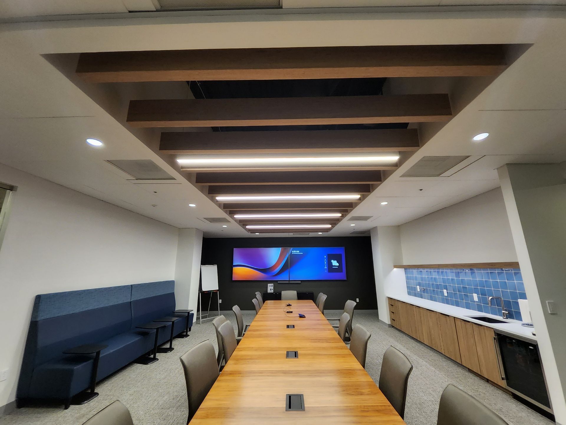 Long conference room with a long wooden table, chairs, and a large screen; ceiling beams and blue and white accents.