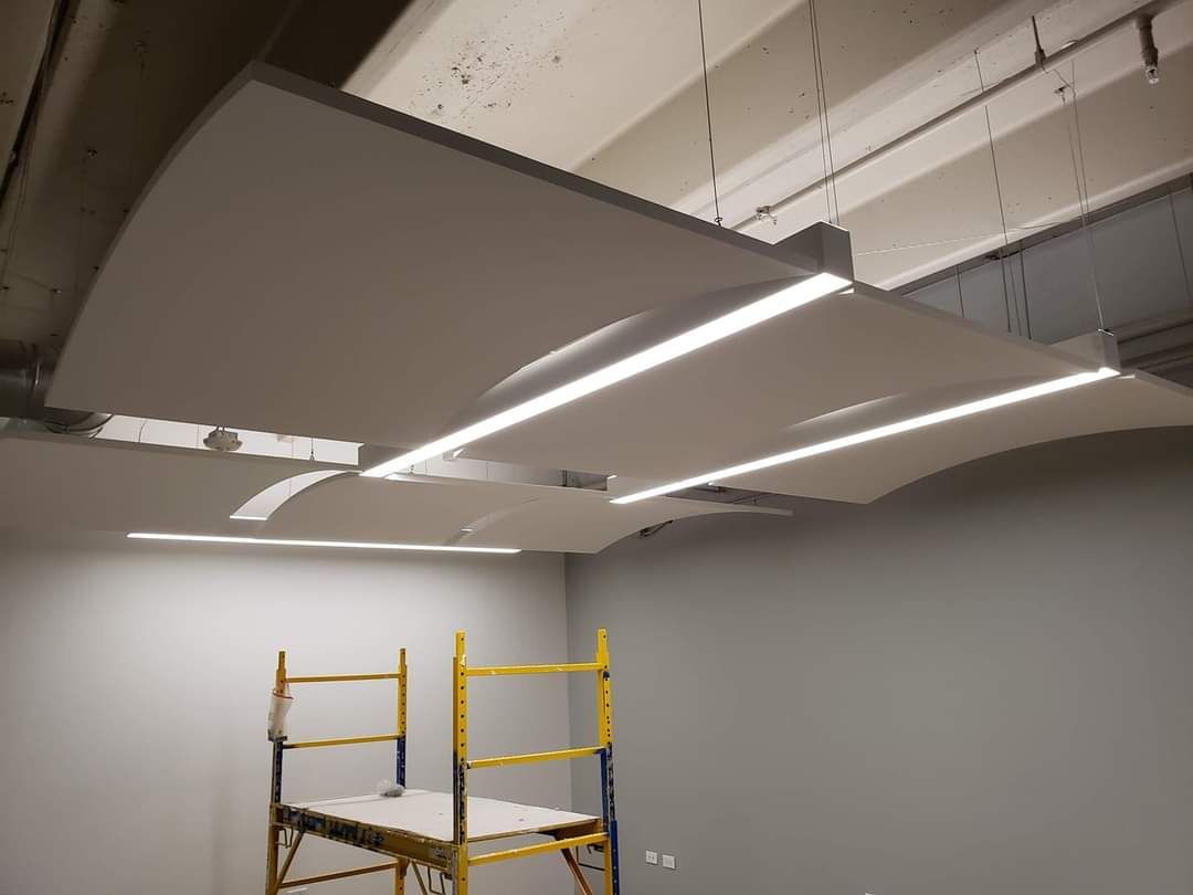 White acoustic panels with integrated lights suspended from a ceiling in a room. A yellow scaffold is below.