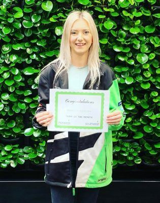 A woman in a green jacket is holding a certificate in front of a green wall.