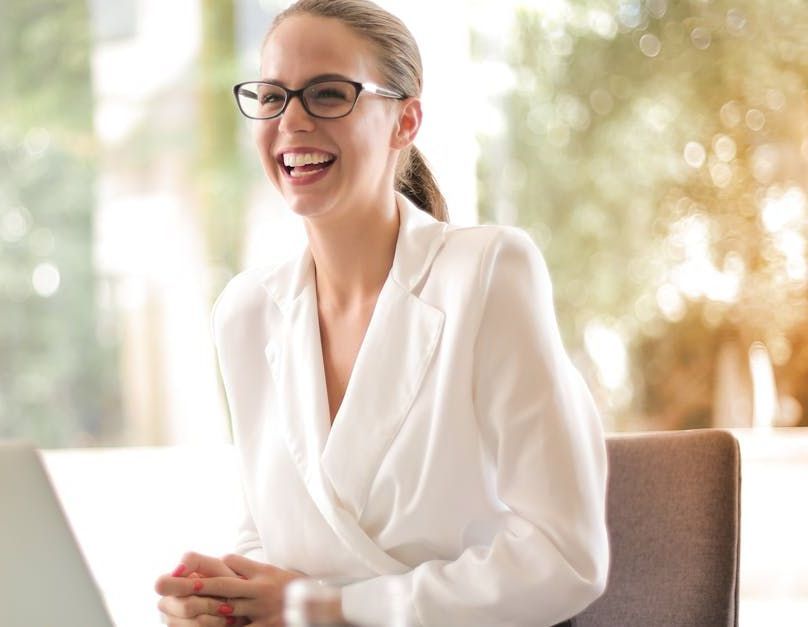 A woman is sitting at a table with a laptop and smiling.