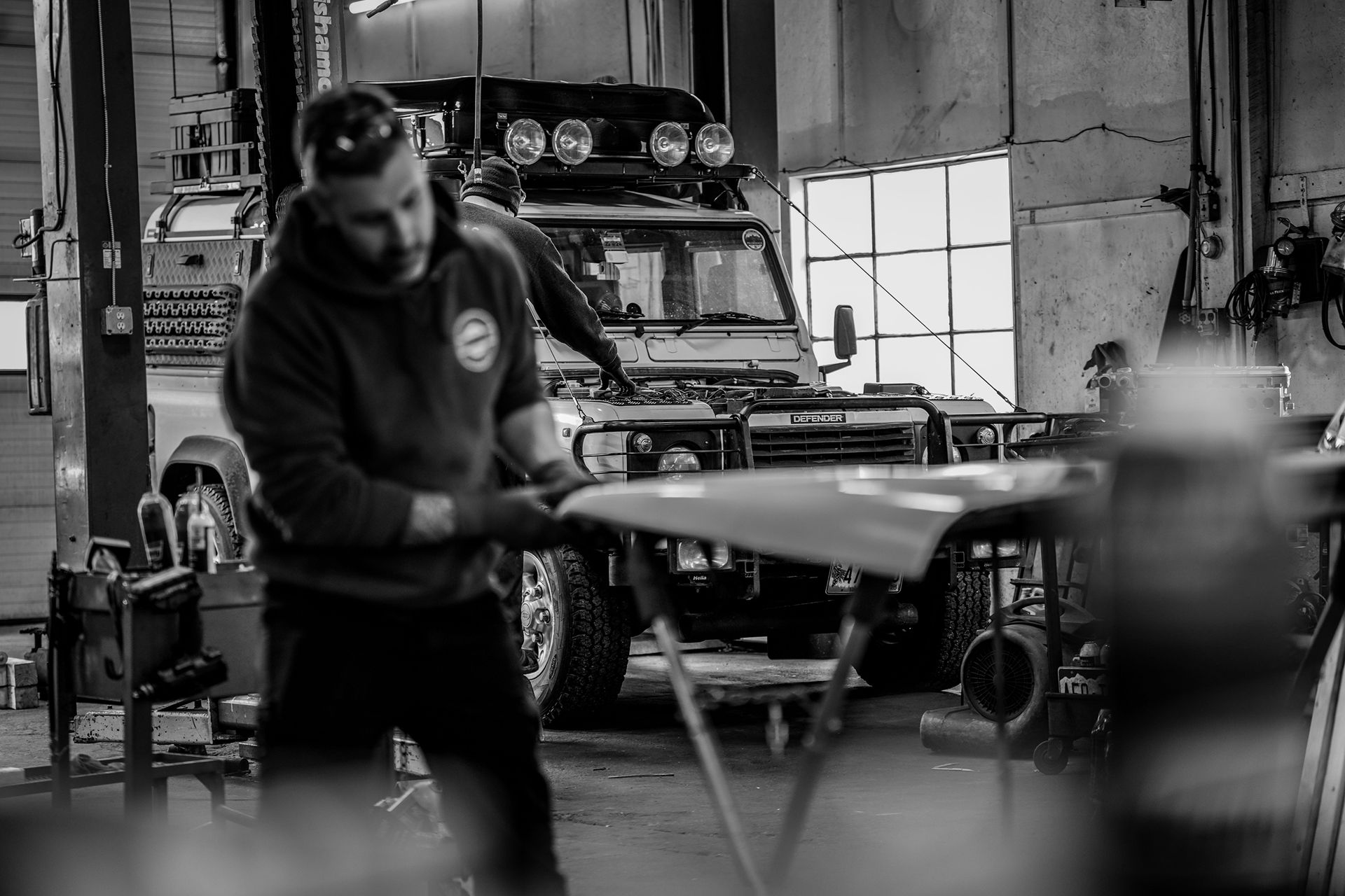 A person working on a car panel in an auto repair shop, with an off-road vehicle visible in the background.