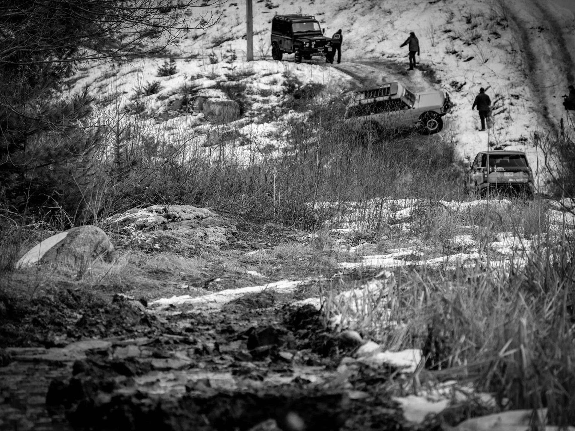Black and white shot of several off-road vehicles navigating a muddy, snowy hillside track with figures standing nearby.