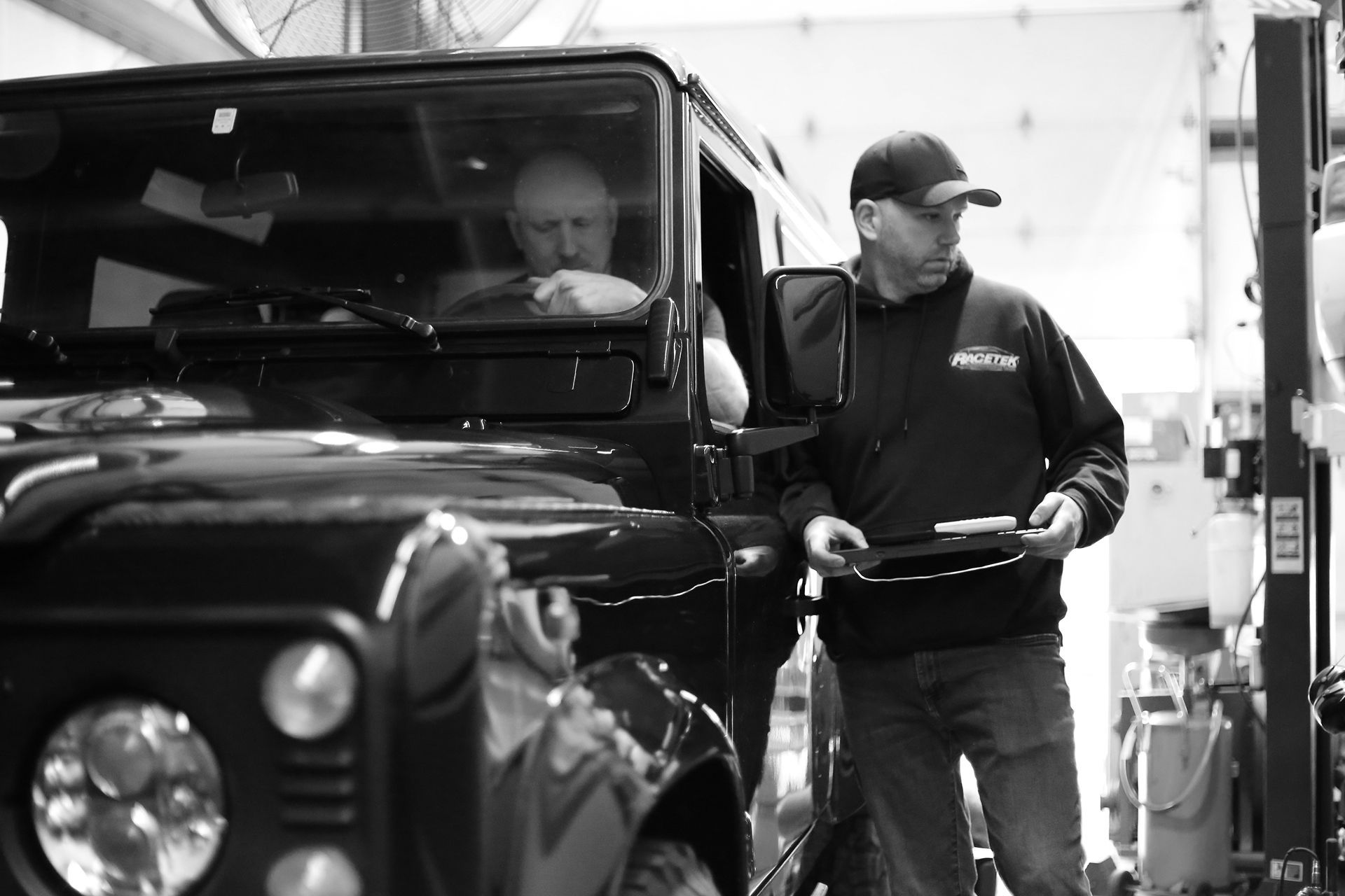 A person in a hoodie holds a clipboard next to a driver in a dark vehicle inside an auto repair shop.
