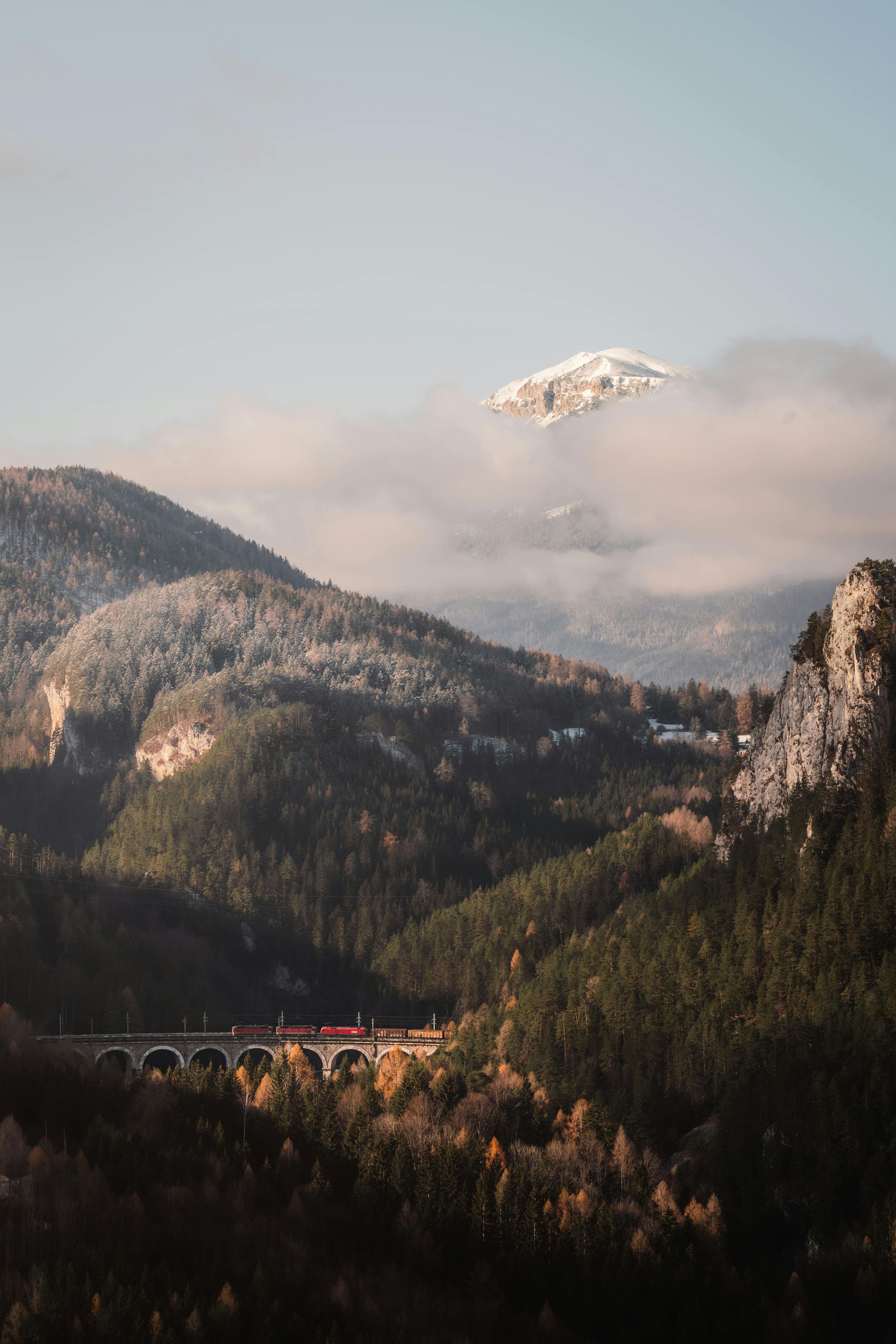 Mountain landscape with a train crossing a bridge, snow-capped peak in the clouds.