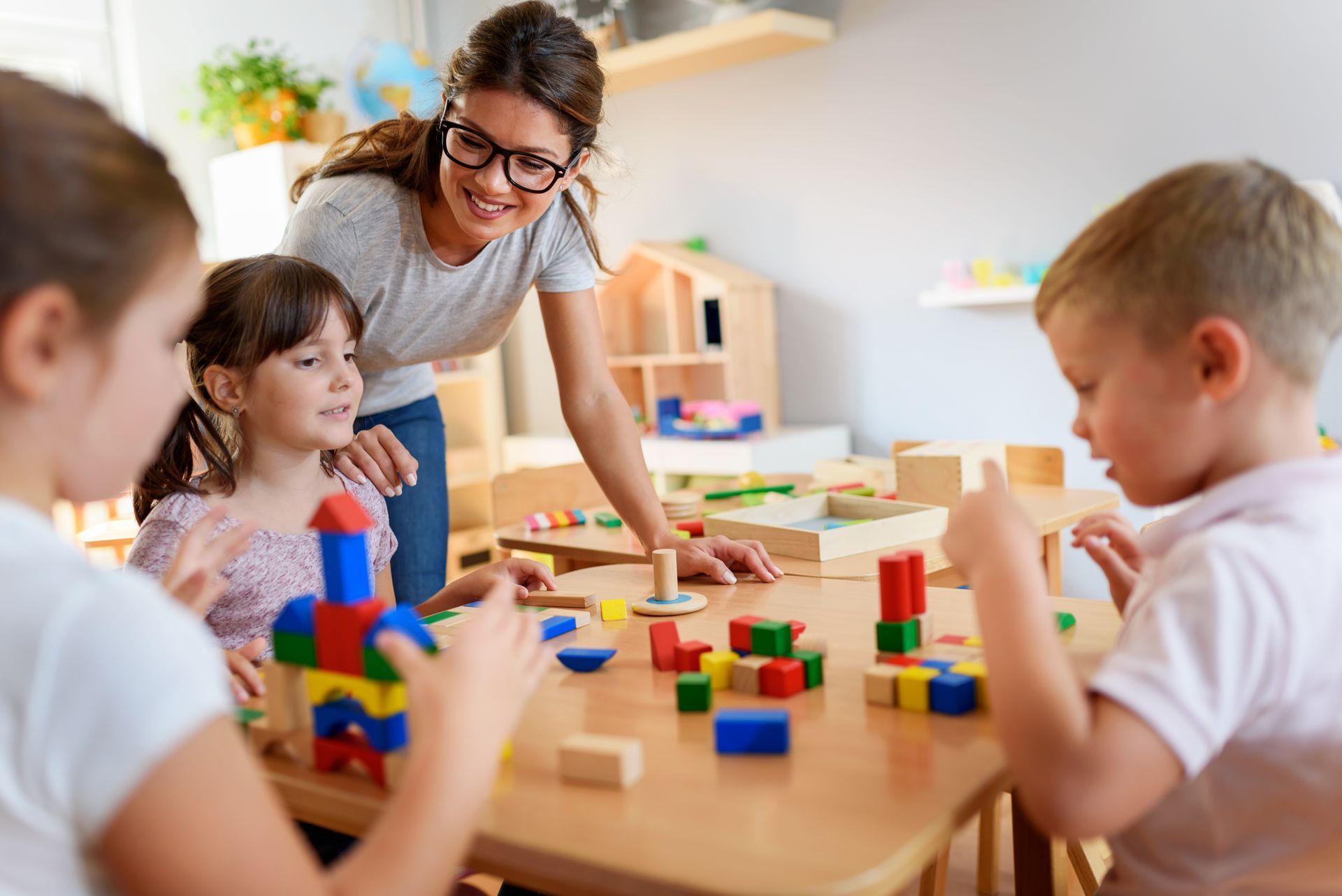 Caregiver supervising young children as they build with colorful wooden blocks at a classroom table.