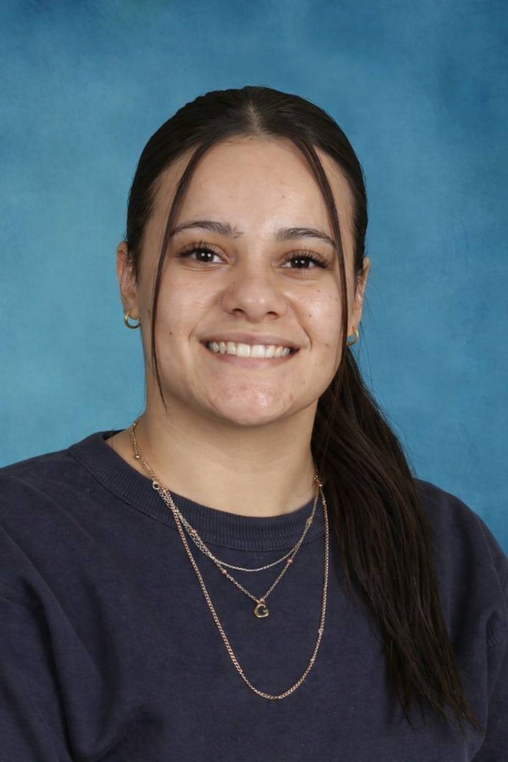 Woman with dark hair smiles, wearing a pink scarf and black top, set against a blue background.