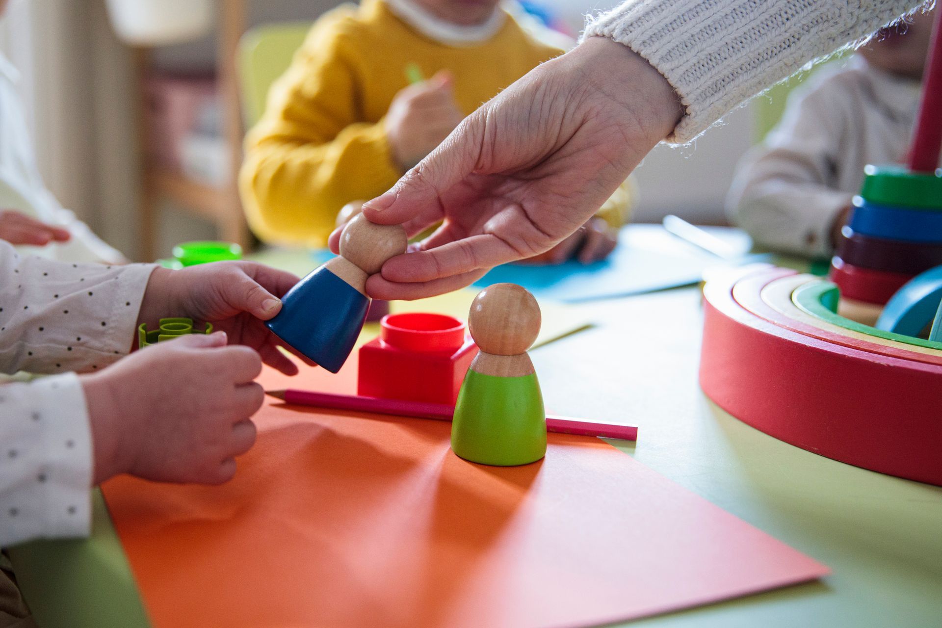 Hand placing a wooden figure on a colorful table with toys and craft materials in a classroom.