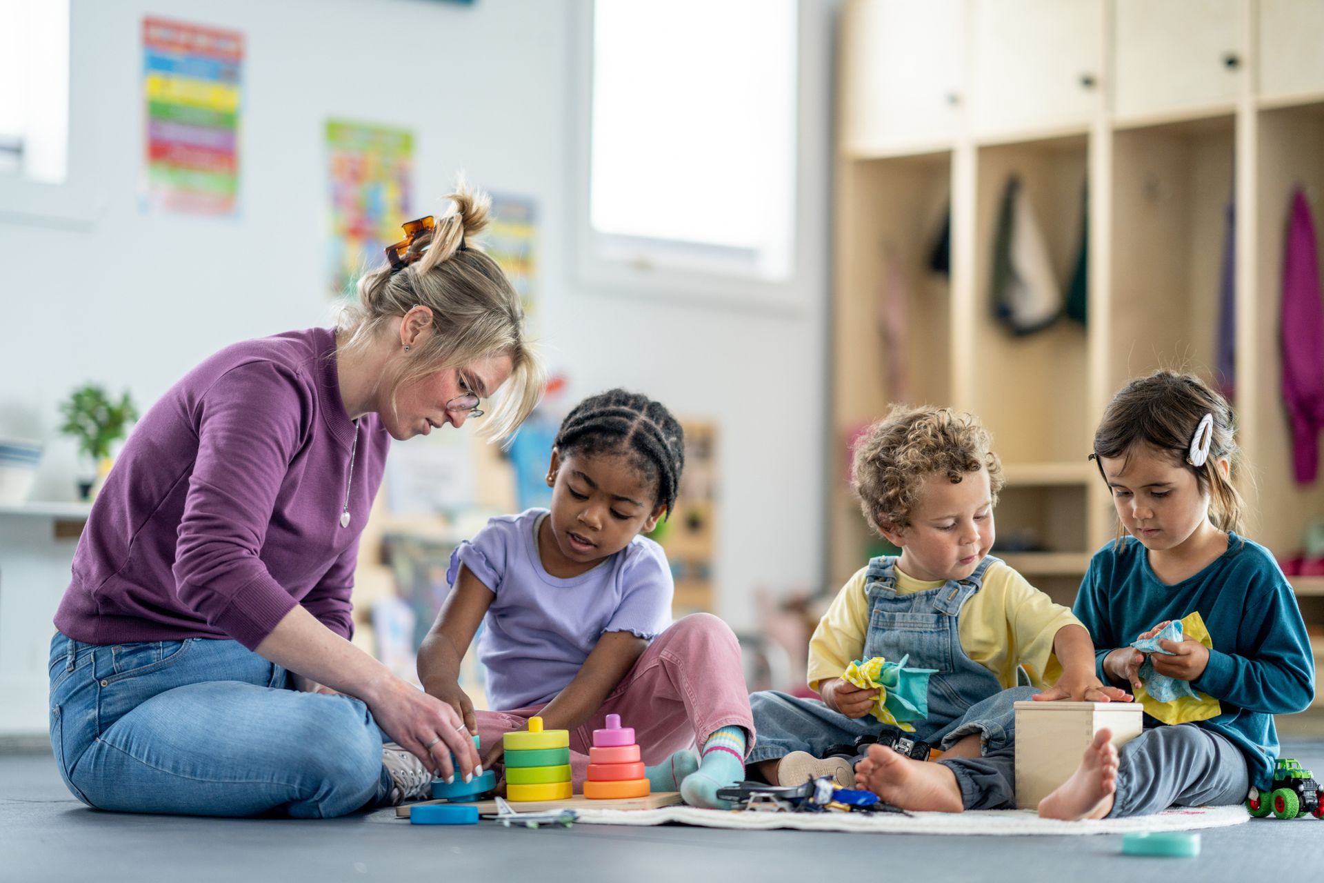Adult sitting on the floor with children playing with colorful stacking toys in a classroom.
