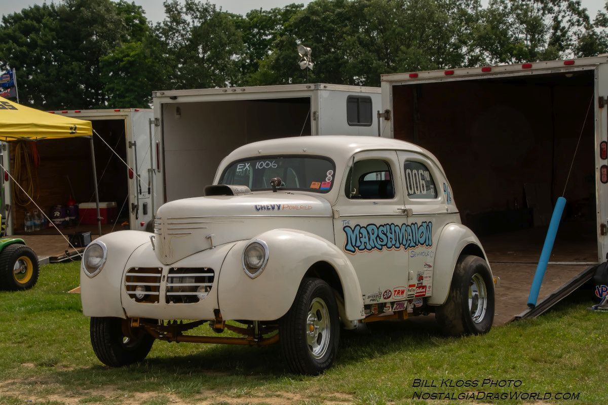 A white car is parked in the grass in front of a trailer.
