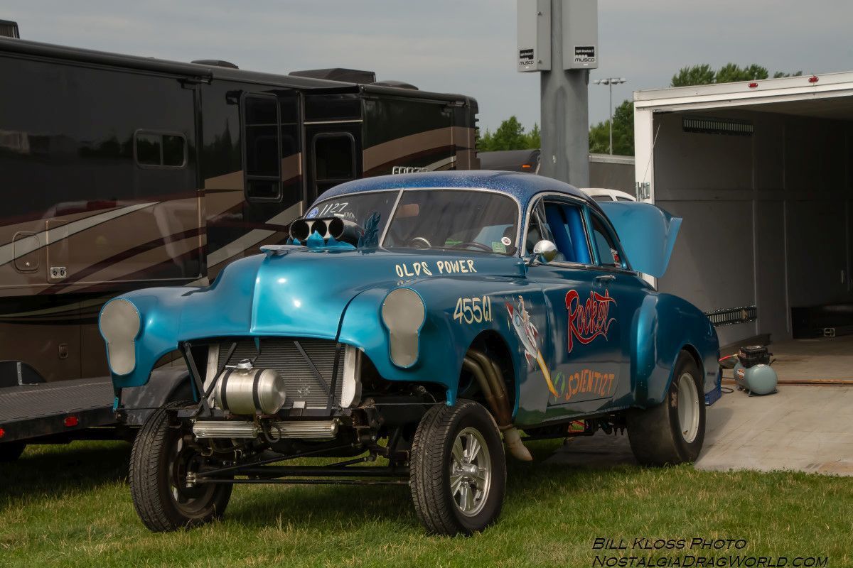 A blue car with the hood up is parked in the grass next to a trailer.