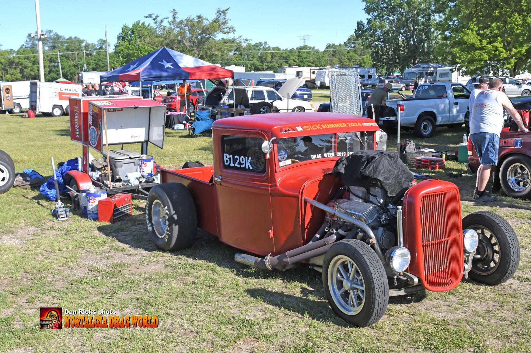 A red hot rod is parked in the grass at a car show