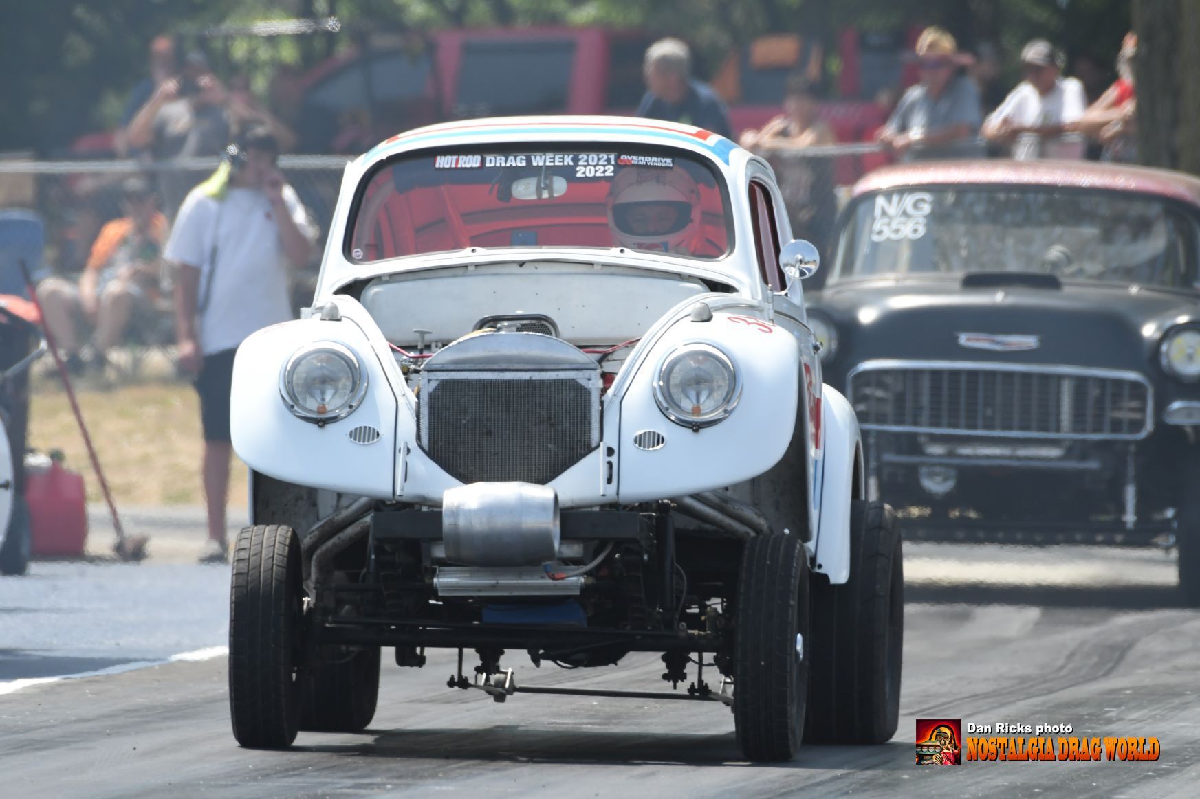 A white car with the hood up is driving down a race track.