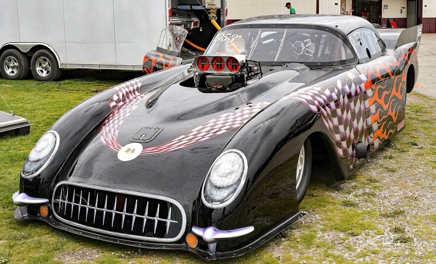 A black race car is parked in a grassy field next to a trailer.