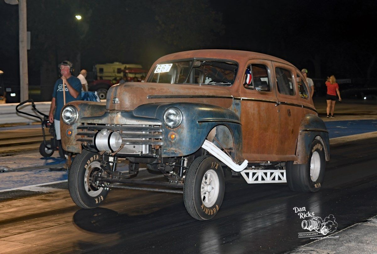 An old rusty car is parked on the side of the road at night.