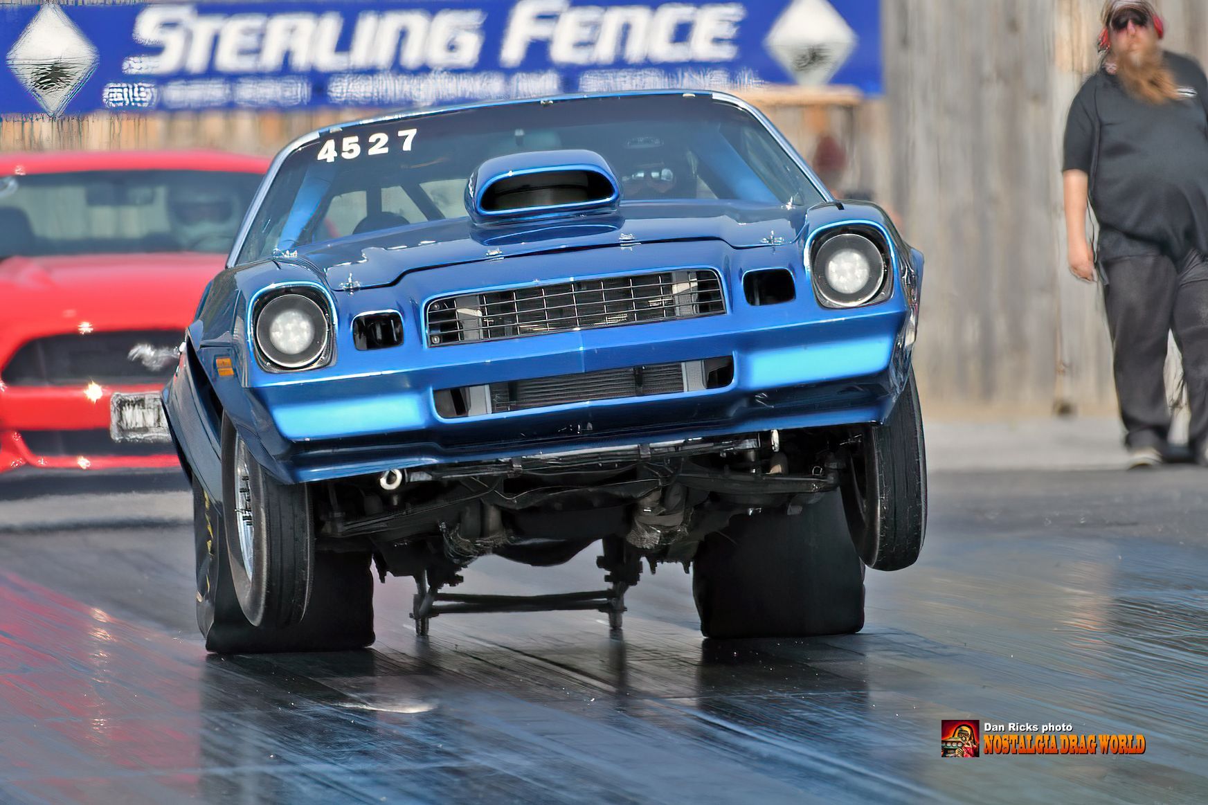 A blue car is driving down a race track in front of a sterling fence sign.