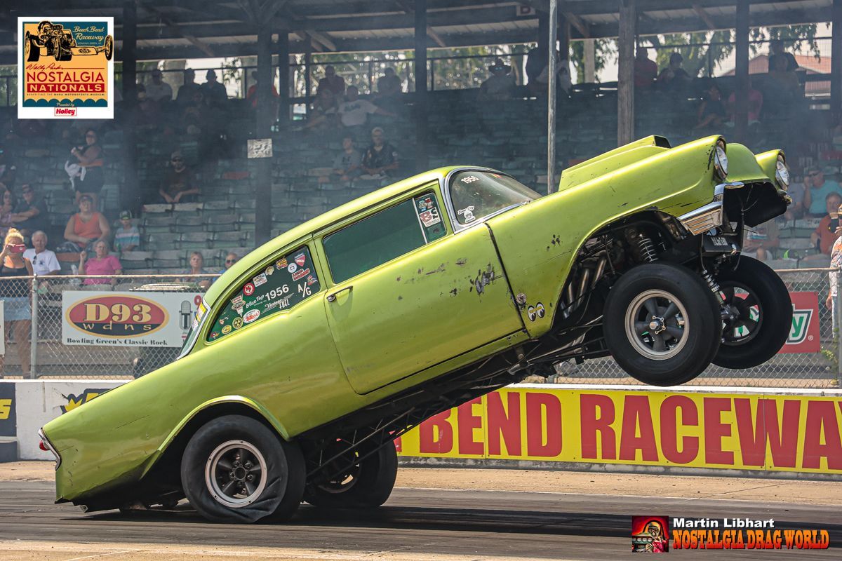 A green car is doing a trick in front of a sign that says bend raceway