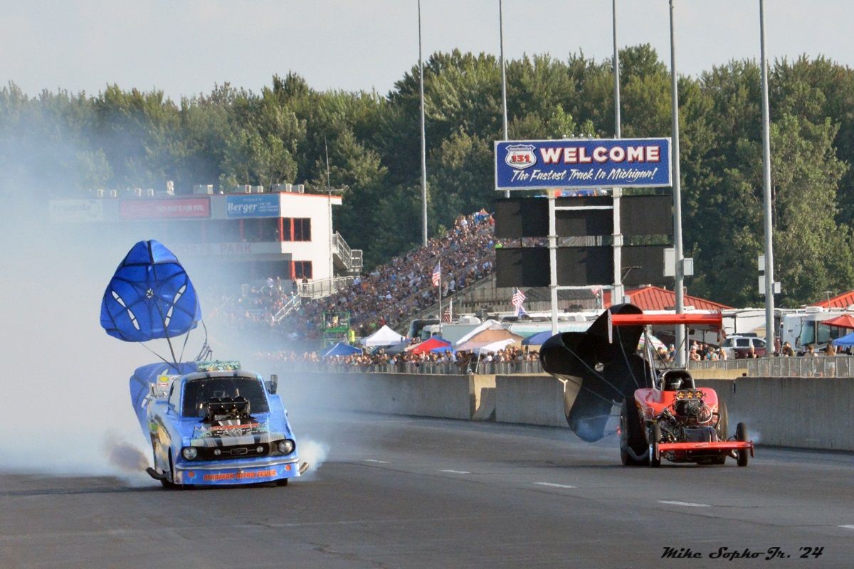 Two drag racing cars on a track with a welcome sign in the background