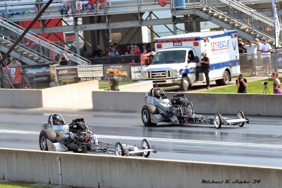 Two drag racing cars are racing on a track with an ambulance in the background.