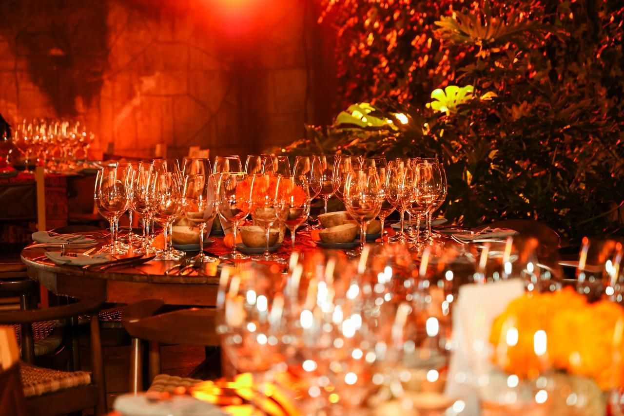 Mesas dispuestas con copas de vino y arreglos florales, cálidamente iluminadas en un ambiente de comedor.