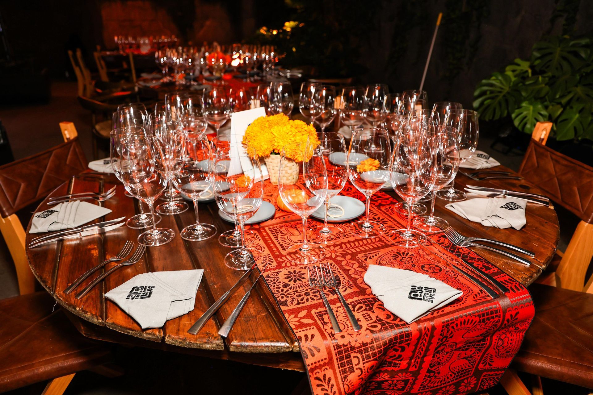 Mesa redonda para cena formal, adornada con cristalería, centro de mesa floral y camino de mesa naranja.