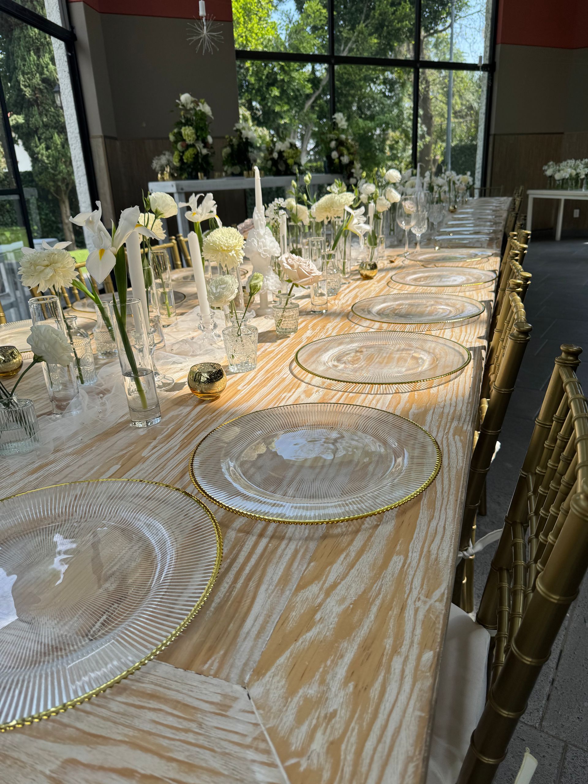 Mesa de comedor larga para evento con decoración en blanco y dorado, grandes ventanales al fondo.