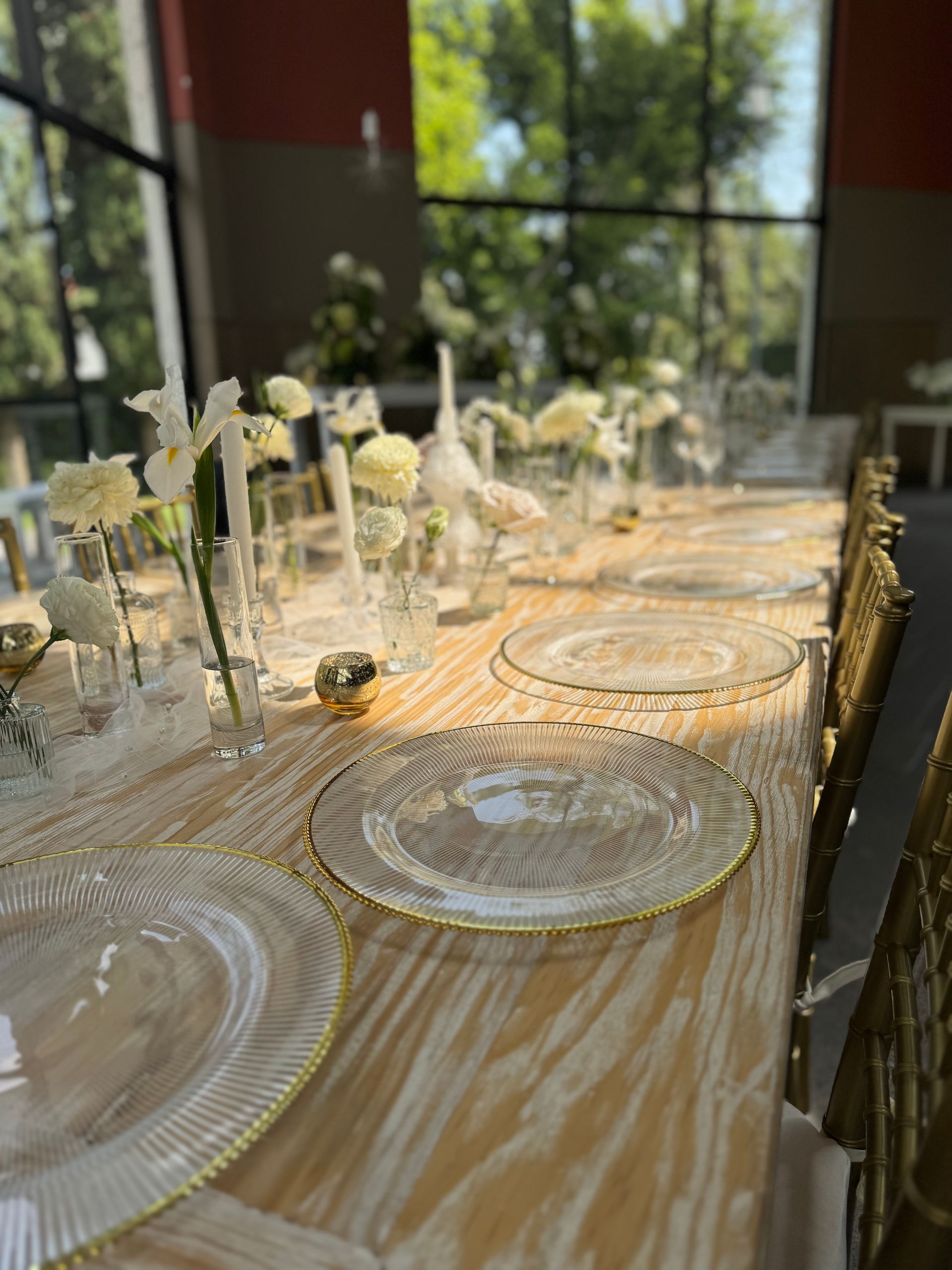 Mesa de comedor larga para un evento formal con detalles dorados, arreglos florales y platos transparentes.