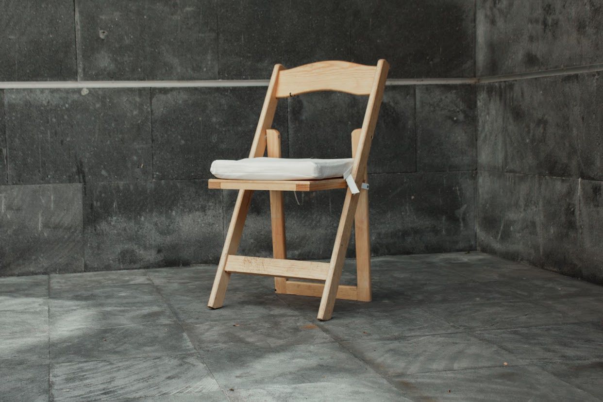 Silla plegable de madera con un cojín blanco contra una pared gris texturizada.