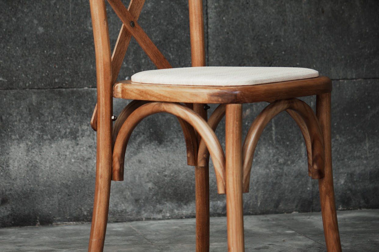 Silla de madera con respaldo cruzado y asiento con cojín color crema, contra una pared oscura y texturizada.