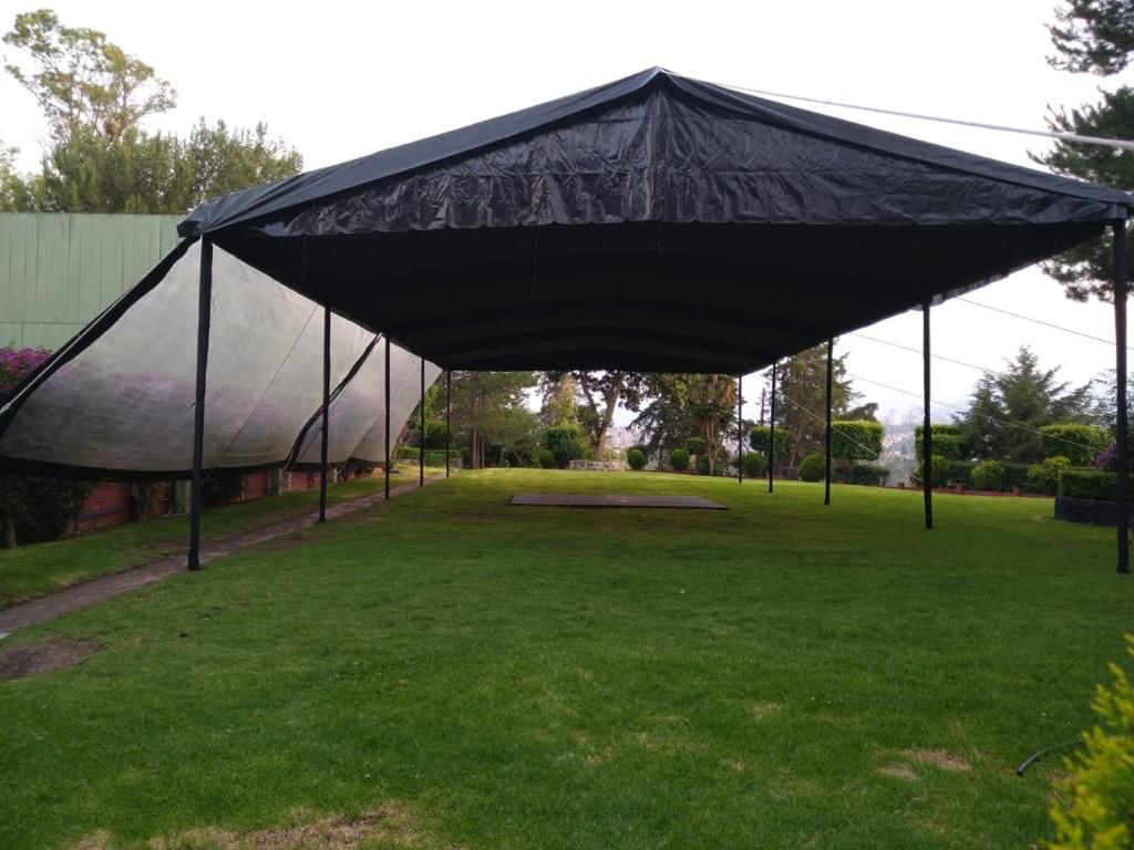 Un dosel negro cubre una zona de césped. Postes metálicos sostienen el techo. Al fondo se ven árboles y follaje verde.