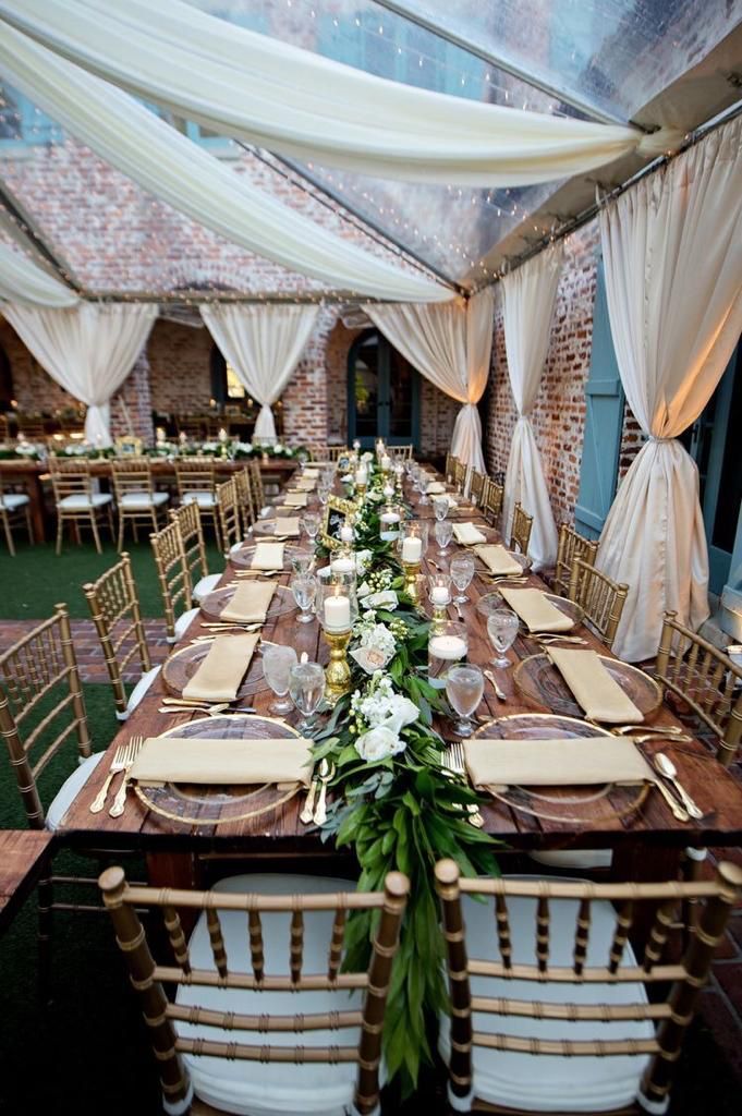 Mesa de comedor larga de madera para un evento formal con sillas doradas y guirnalda floral; ambiente al aire libre con carpa.