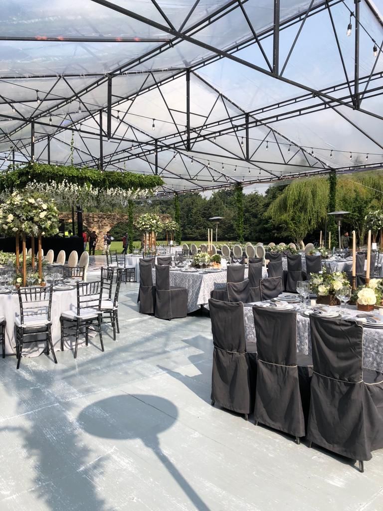 Montaje de recepción de boda al aire libre con mesas, sillas y arreglos florales bajo una estructura de techo transparente.