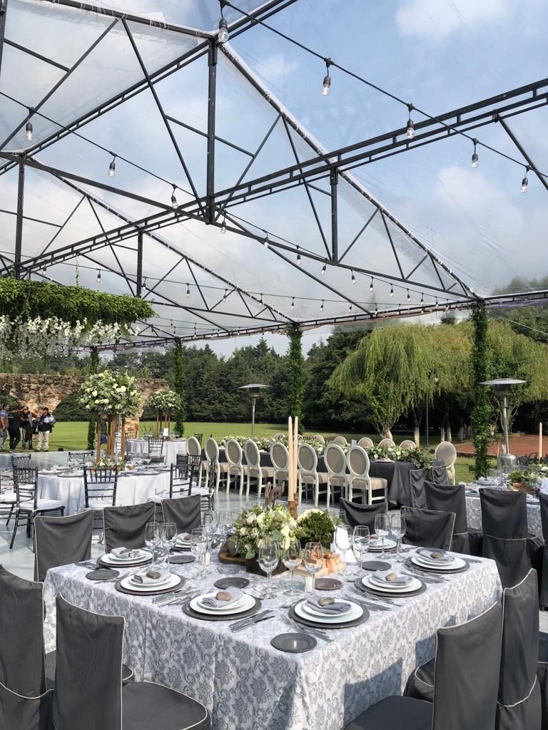 Recepción de boda bajo un dosel transparente. Mesas decoradas en gris y blanco, con los invitados al fondo.