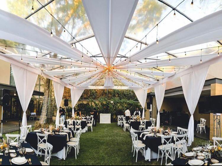 Recepción de boda bajo una carpa transparente con cortinas blancas, mesas con manteles negros y luces de cadena.