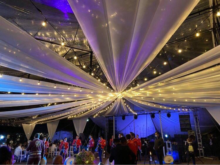 Lugar para eventos con cortinas blancas, luces de cadena y un escenario con gente.