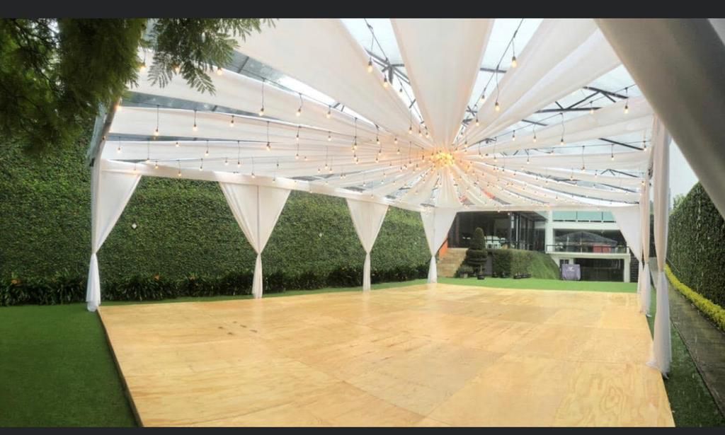 Pista de baile de madera bajo un dosel cubierto con luces de cadena, rodeada de setos y un edificio de cristal.