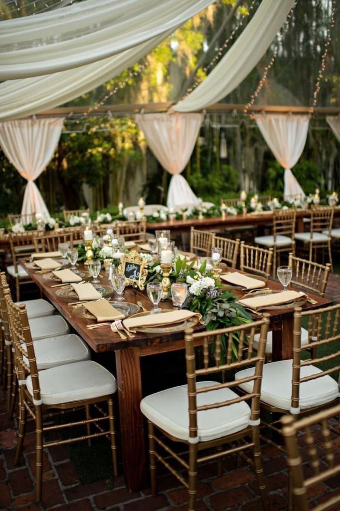 Montaje de la recepción de boda: largas mesas de madera, sillas doradas con cojines blancos, telas drapeadas, vegetación y velas.