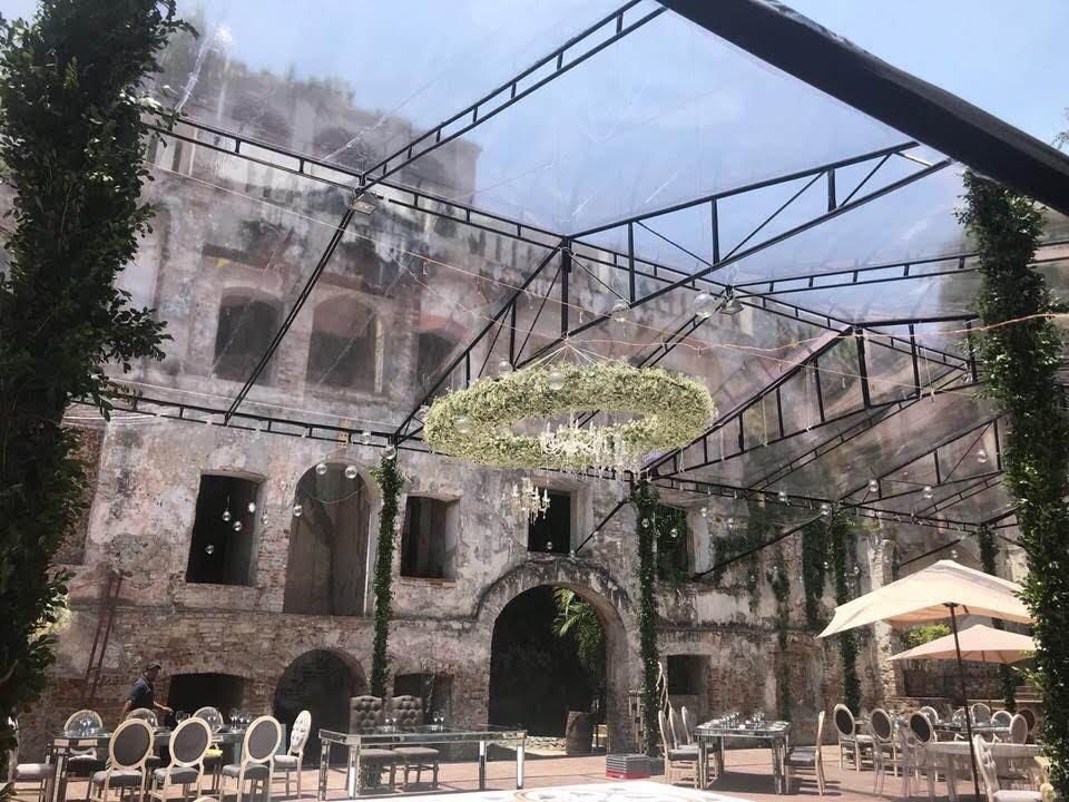 Ceremonia de boda en un antiguo edificio de ladrillo, cubierta por una carpa transparente. Una lámpara de araña con flores cuelga sobre los asientos.