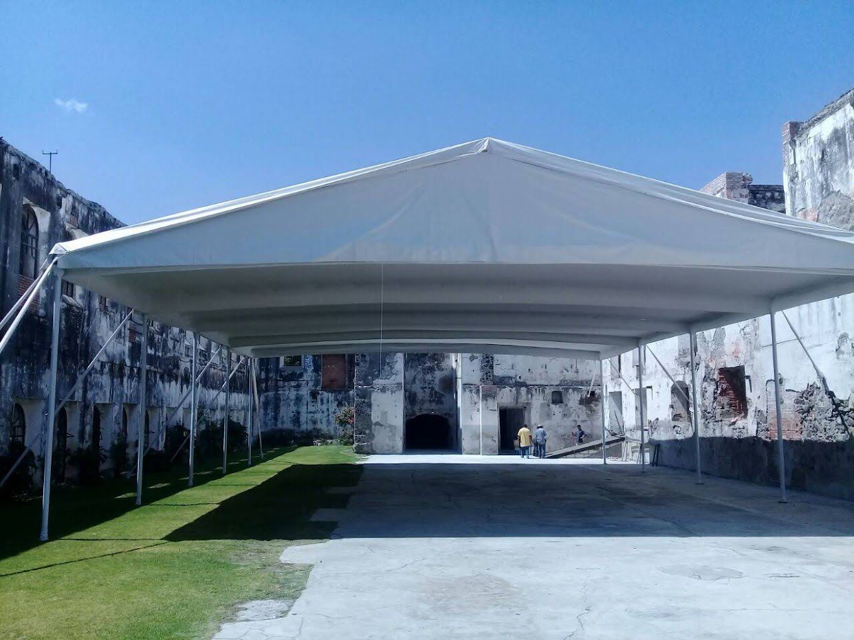 Un dosel blanco sobre un camino de hormigón, flanqueado por un antiguo edificio de piedra erosionada. Día soleado.