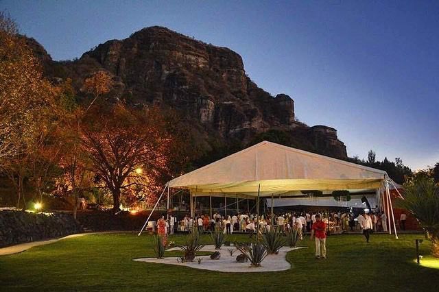 Evento al aire libre bajo una carpa blanca en un césped con invitados y fondo de montaña al anochecer.