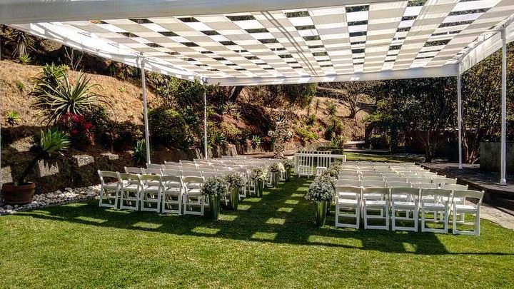 Instalación de ceremonia de boda al aire libre con sillas blancas sobre césped verde bajo un dosel a cuadros.