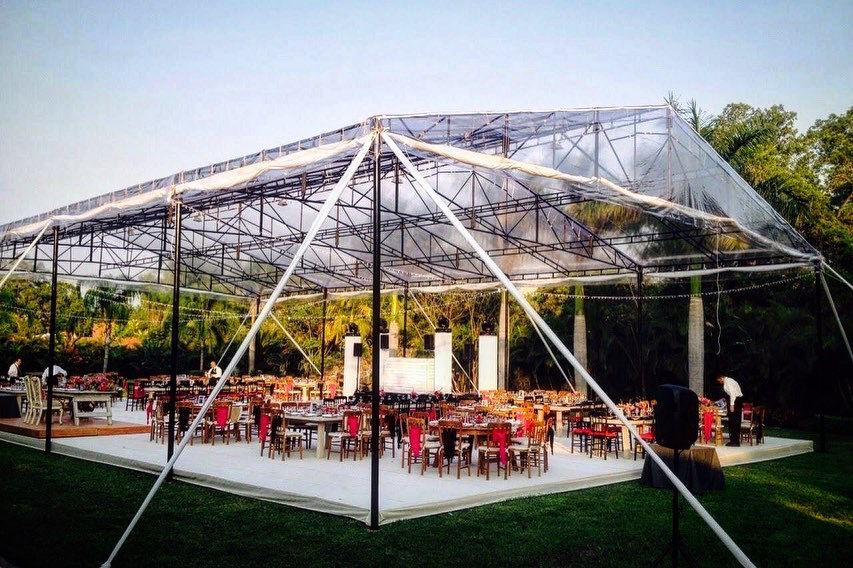 Carpa con techo transparente sobre un área de comedor con mesas y sillas, colocada sobre una plataforma blanca, al aire libre sobre césped.