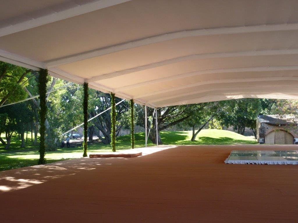 Área exterior cubierta con piso marrón, soportes blancos y vista a árboles y césped.