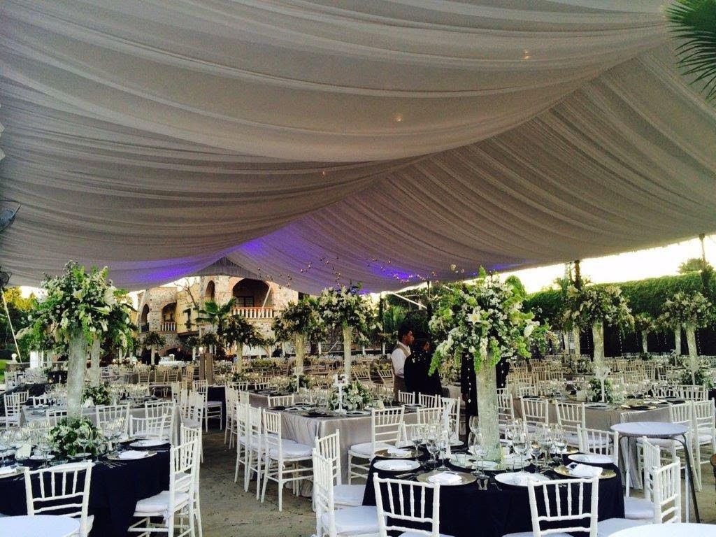 Recepción de boda bajo un dosel blanco drapeado, con mesas con sillas blancas y centros de mesa florales.