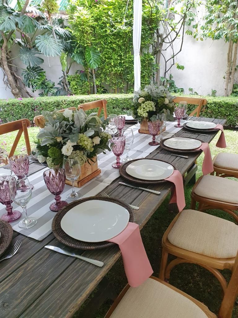 Mesa larga de madera para evento al aire libre, con centros de mesa florales, platos y cristalería rosa.