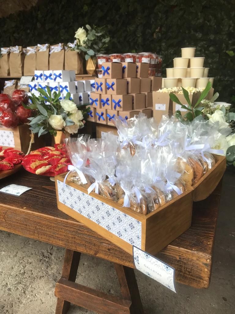 Mesa de madera que exhibe productos horneados, golosinas y recuerdos en un evento, con arreglos florales y cajas de regalo.