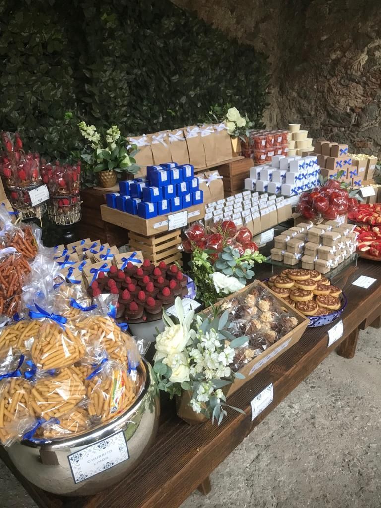 Una mesa de madera muestra una variedad de golosinas y flores, incluidas galletas, bayas y regalos empaquetados, al aire libre.