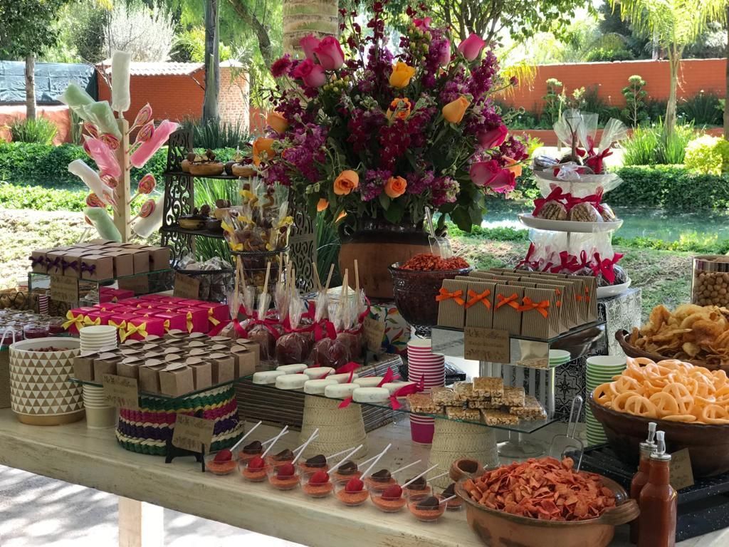 Mesa de postres con diversos dulces, flores y snacks al aire libre.