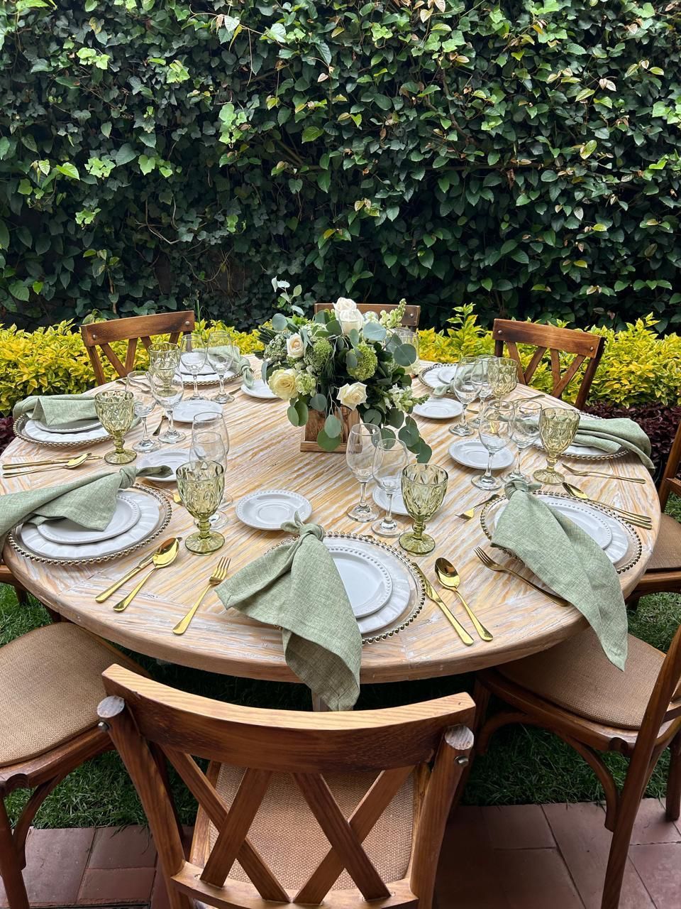 Mesa redonda dispuesta para comer al aire libre, con centro floral y cubiertos de platos, servilletas y vasos.