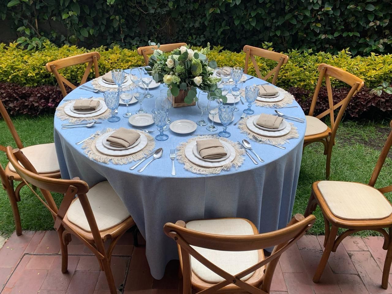 Mesa redonda preparada para comer con mantel celeste, sillas de madera y centro de mesa floral al aire libre.