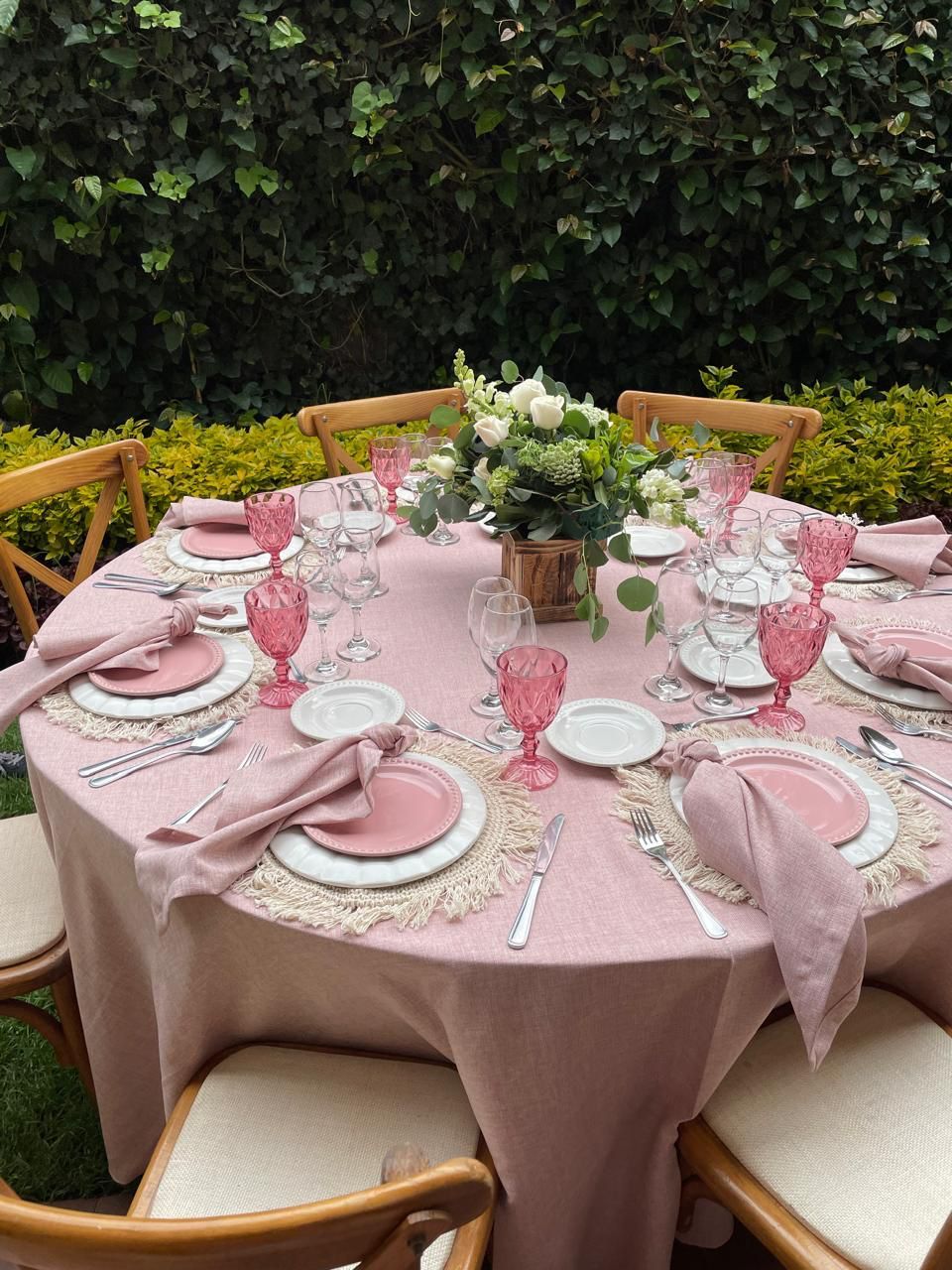 Mesa redonda para comer al aire libre, con mantelería rosa. Incluye cubiertos, cristalería y un centro de mesa floral.
